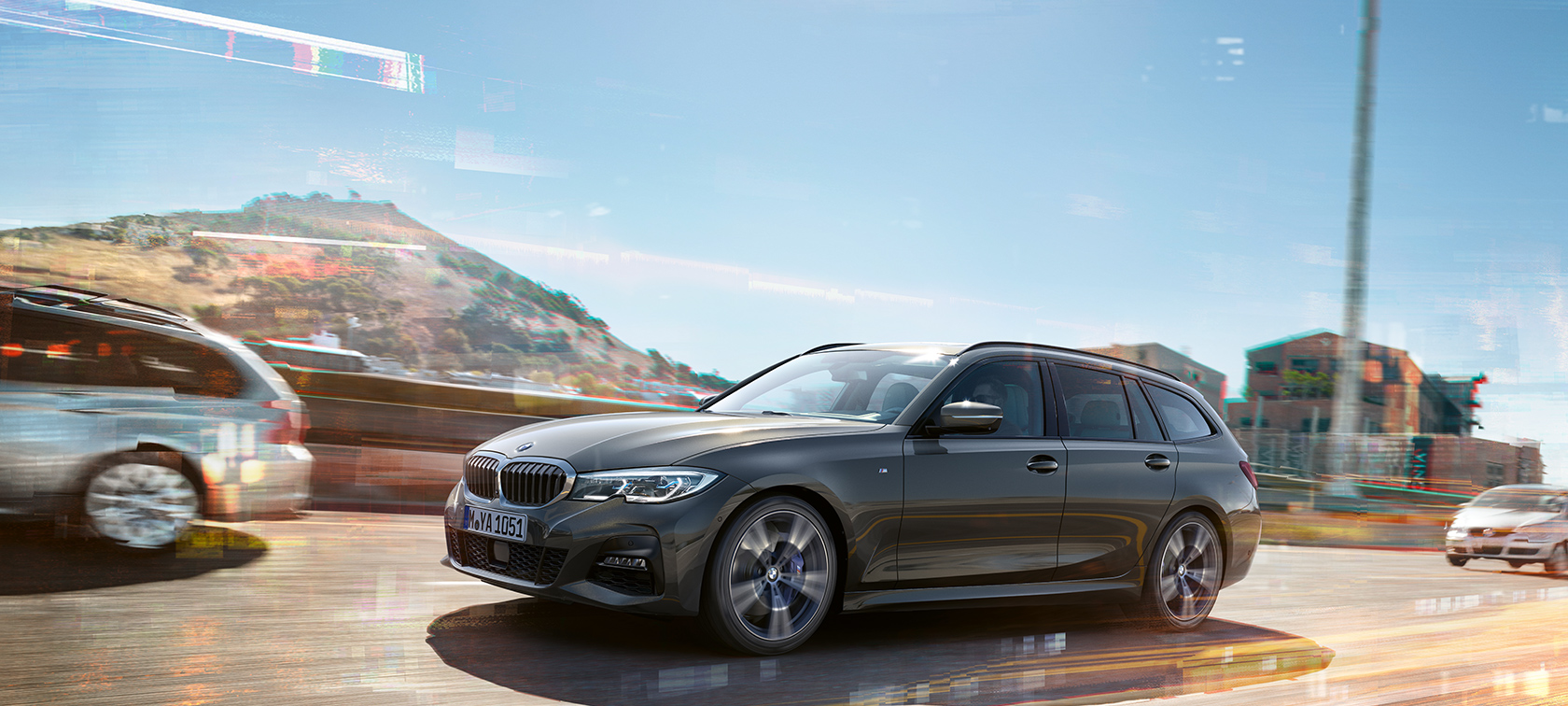 BMW 3 Series Touring (G21),  Mineral Grey metallic, ripresa laterale in movimento con case e montagne sullo sfondo